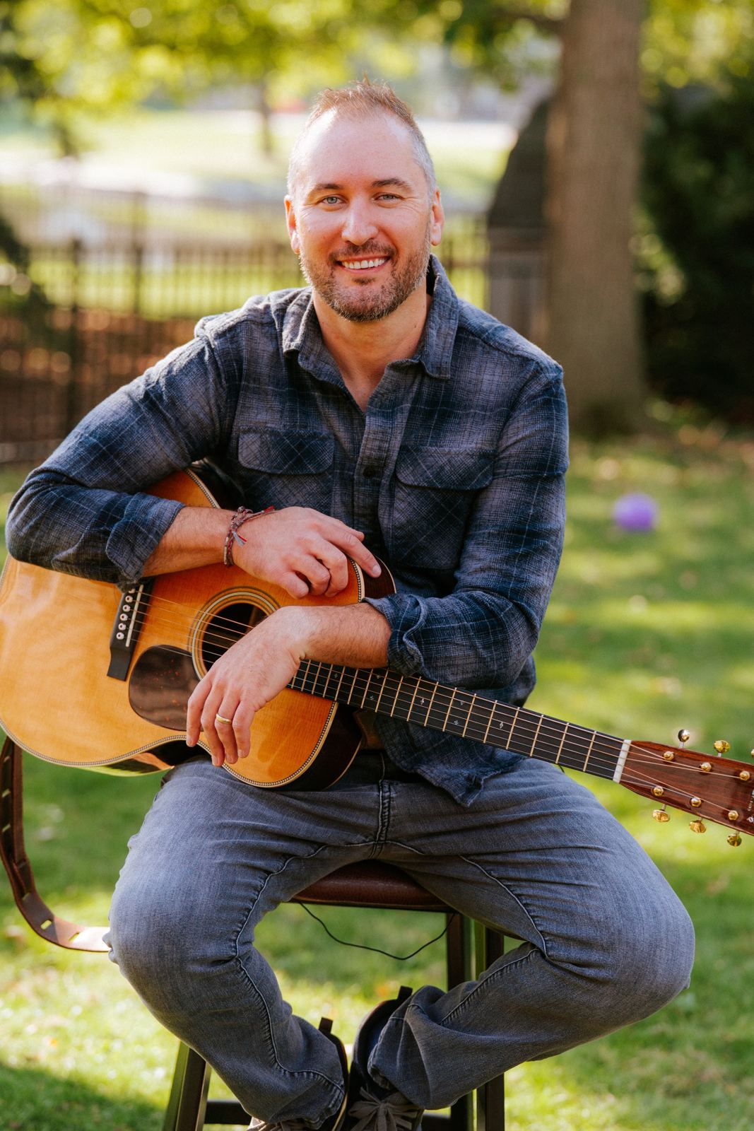 John Crean smiling, seated outdoors with his acoustic guitar